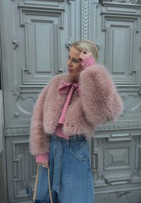 Femme portant une veste rose duveteuse et un chemisier avec un nœud, un jean bleu taille haute, tenant un sac à main bleu marine, debout près d'une porte grise décorative.