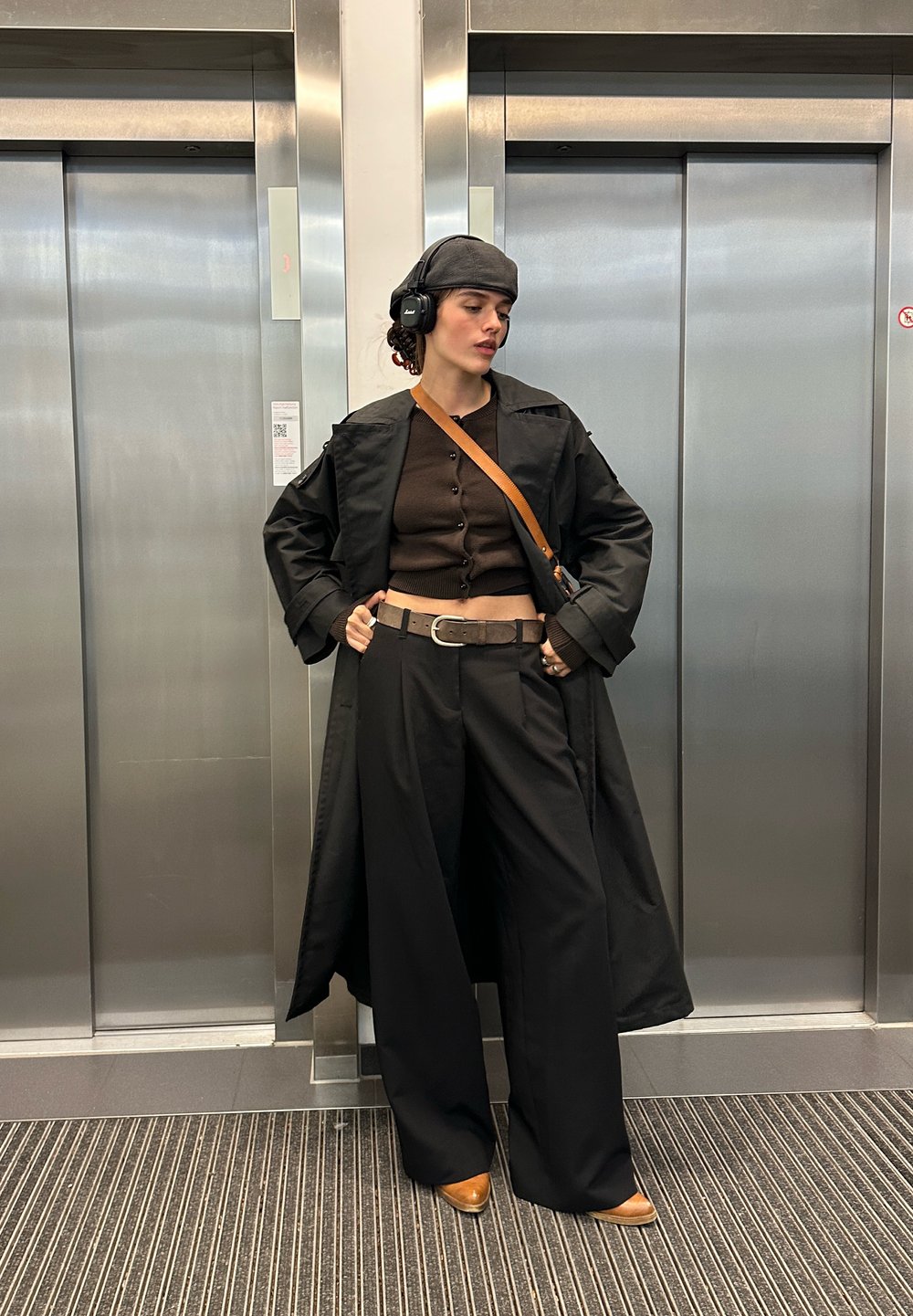 Čierny dlhý kabát cez hnedý crop top, široké čierne nohavice a hnedé topánky. Doplnky zahŕňajú šedú čiapku a veľké čierne slúchadlá.