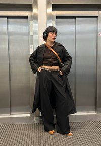 Čierny dlhý kabát cez hnedý crop top, široké čierne nohavice a hnedé topánky. Doplnky zahŕňajú šedú čiapku a veľké čierne slúchadlá.