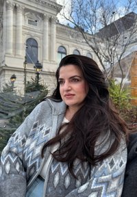 Grå cardigan med geometriske blå og hvite mønstre, med glidelåslukking og strukturert strikk. Modellen har langt, mørkt hår og befinner seg utendørs.