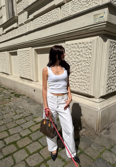 Top blanco de tirantes combinado con jeans blancos de cintura alta. Lleva un bolso de mano marrón con monograma y correas rojas brillantes, además de zapatos negros de punta.