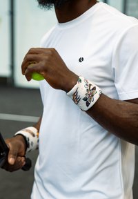 Valge sportlik särk, dekoratiivsed triibulised randmerihmad erkade mustritega, hoides rohelist tennispalli. Lähedalt pildistatud käed ja aksessuaarid.