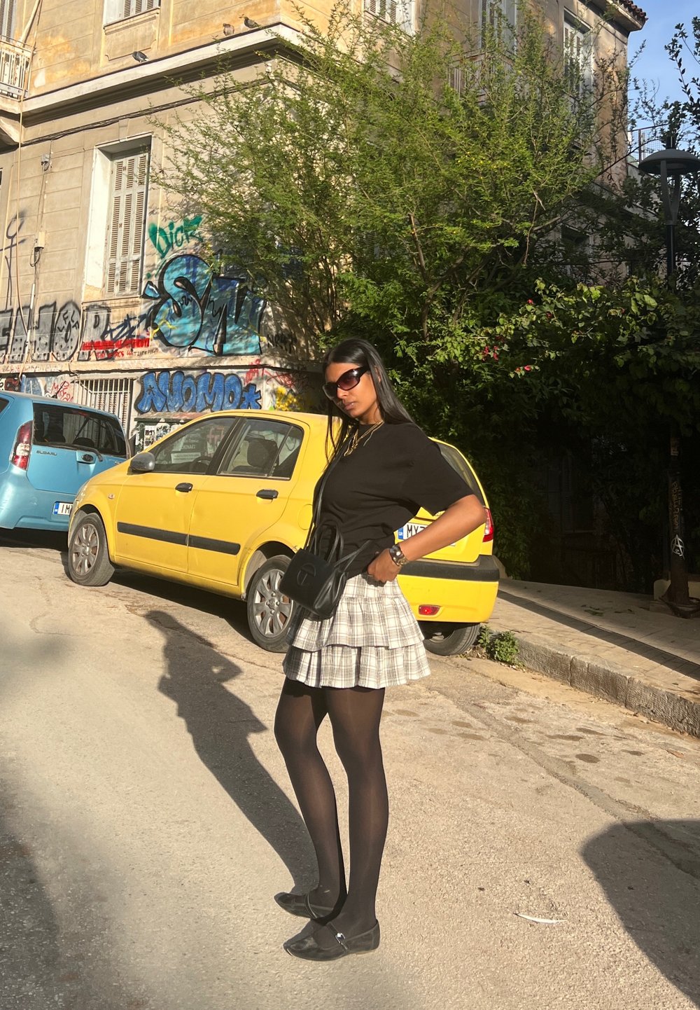 Μαύρο κοντομάνικο τοπ, φούστα με καρό και πτυχώσεις, μαύρες καλσόν, μαύρες μπαλαρίνες. Μικρή μαύρη τσάντα, γυαλιά ηλίου. Αστική σκηνή με γκράφιτι.