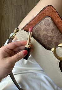 Läppstift med en rosapink nyans i ett guld- och svart rör, hålls ovanför en beige och brun texturerad väska. Vit tyg under.