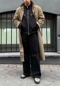 Άτομο που φοράει ένα μπεζ παλτό τύπου τρεντς πάνω από μια μαύρη τζάκετ με φερμουάρ και φαρδιά μαύρα παντελόνια, στέκεται με τα χέρια στις τσέπες σε πλακόστρωτο πεζοδρόμιο.