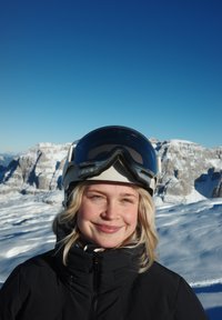 Casque de ski blanc avec visière teintée, veste noire zippée, cheveux blonds naturels, arrière-plan de montagne enneigée, ciel bleu clair.