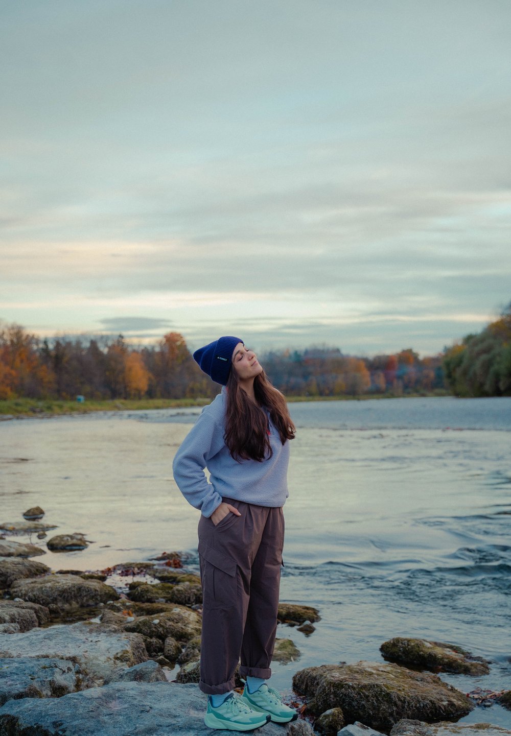 Mikina z fleecu světle šedé barvy, volné hnědé cargo kalhoty a tyrkysové tenisky. Navy modrá čepice doplňuje ležérní venkovní vzhled u vody.