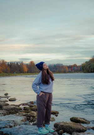 Sudadera de forro polar en gris claro, pantalones cargo marrones sueltos y zapatillas deportivas en color teal. Un gorro de lana azul marino completa el look casual al aire libre cerca del agua.