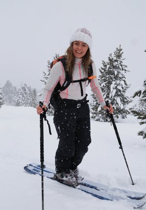 Mujer sonriendo mientras esquía en un paisaje nevado, con un gorro de punto blanco, chaqueta rosa y blanca, pantalones negros y llevando una mochila.