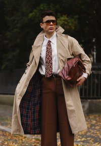 Bež trench coat, millel vooderdus, valge särk, mustriga burgundia tie, pruunid püksid ja pruun nahkrahakott. Päikeseprillid täiendavad välimust.