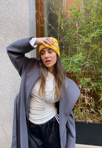 Manteau oversized gris, haut ajusté blanc, pantalon de sport noir avec des rayures, bonnet en tricot jaune à motif et colliers superposés. Arrière-plan avec du bambou.