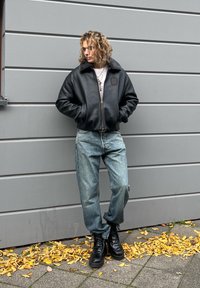 Mustast bomberjakk hõlmiknahkmööbliga kraega, tõmblukuga ees ja logomärgiga. Hele sinine lahtine teksapüksid ja mustad paeltega saapad täiendavad outfiti.