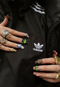 Casaco preto com o logótipo branco da Adidas, apresentando designs de unhas intrincados e coloridos, anéis e um relógio prateado nas mãos.