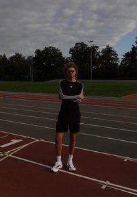 Camisola atlética de manga longa preta com riscas brancas, calções pretos, ténis brancos e óculos de sol, em pé numa pista de corrida.