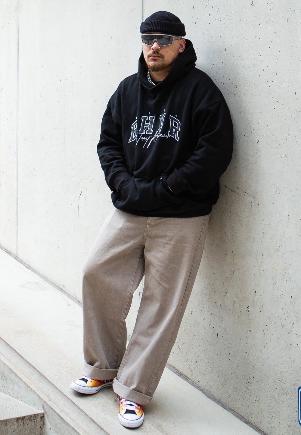 Black hoodie with white logo, beige wide-leg pants, and colorful sneakers. Accessories include gray sunglasses and a black beanie.
