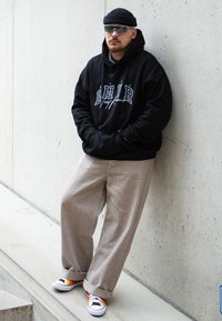 Black hoodie with white logo, beige wide-leg pants, and colorful sneakers. Accessories include gray sunglasses and a black beanie.