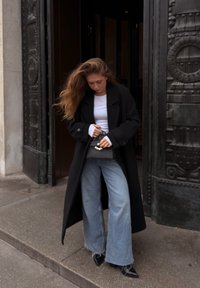 Svart lang frakk, hvit topp og vide blå jeans. Holder en liten svart veske. Glatte teksturer med kontrasterende farger. Står på en steinbelagt overflate.