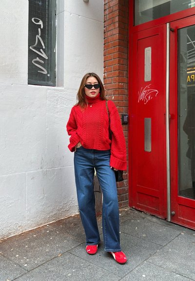 Suéter de punto rojo con cuello alto y patrón de trenzas, combinado con jeans de pierna ancha azules y zapatos rojos de slip-on; de pie junto a una puerta roja.