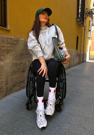Sudadera gris, mallas negras, calcetines blancos con acentos rojos y zapatillas plateadas. Persona en una silla de ruedas con una bolsa verde de patrones, situada frente a una pared amarilla.