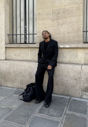 Traje negro, camisa negra y pantalones de pierna ancha. Los accesorios incluyen anillos y gafas transparentes. Una bolsa de viaje negra reposa en el suelo.