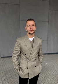 Dubbelzijdige beige blazer met verticale strepen, gecombineerd met een taupe trui en zwarte broek. Grijze textuur achtergrond en trottoir.