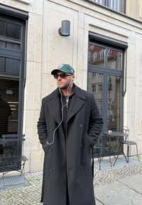 Zwarte dubbelrijige jas over een donker truitje, groene pet, zwarte zonnebril en oortjes, staande voor een stenen muur en buitenzitplaatsen.