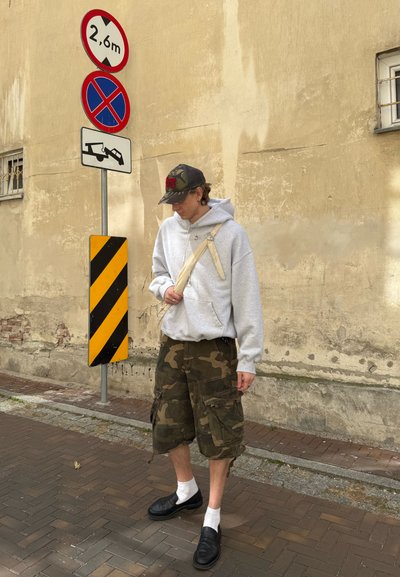 Sudadera gris con capucha, pantalones cortos cargo de camuflaje, calcetines blancos, zapatos negros sin cordones. De pie cerca de señales de tráfico contra una pared texturizada.