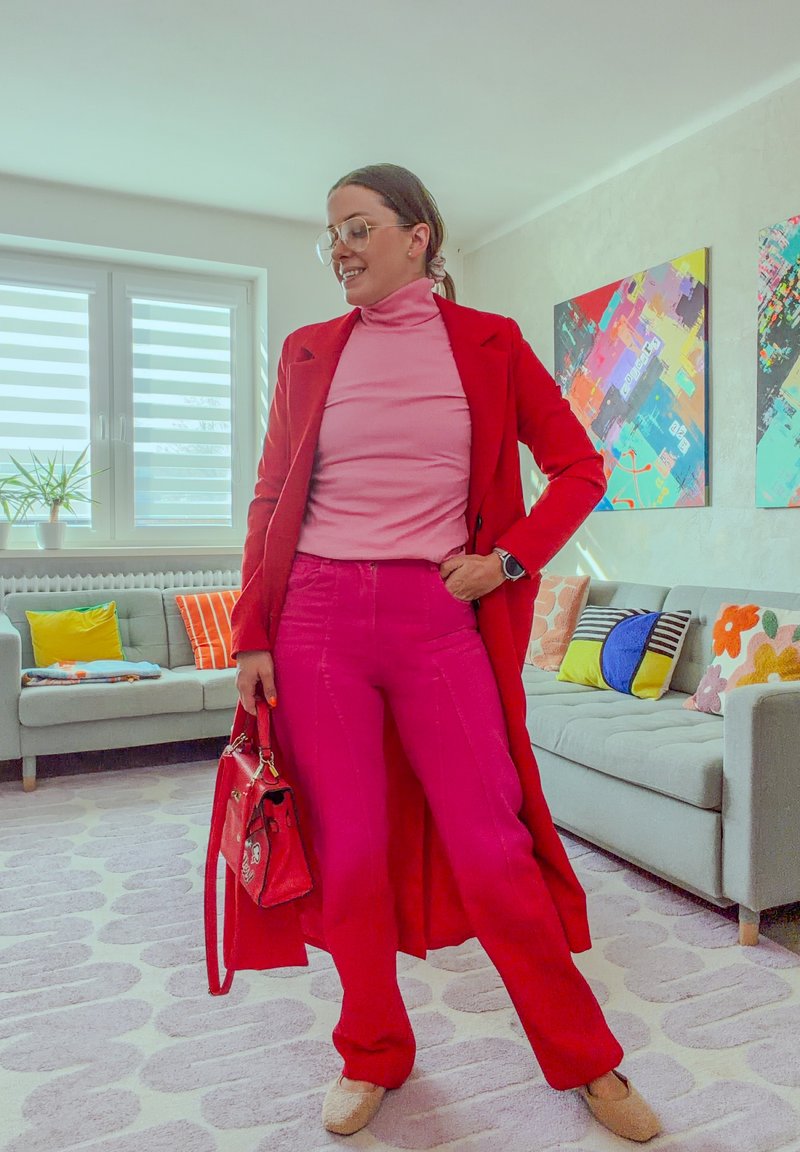 Cappotto lungo rosso, dolcevita rosa e pantaloni fucsia vivaci. Accessoriato con una piccola borsa rossa. Stanza luminosa con cuscini colorati e opere d'arte.
