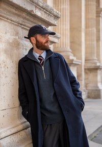 Casaco de lã azul-marinho com lapelas, suéter escuro com fecho, camisa de vestir azul-claro, gravata com riscas bordeaux e boné de beisebol azul-marinho. Fachada de betão ao fundo.