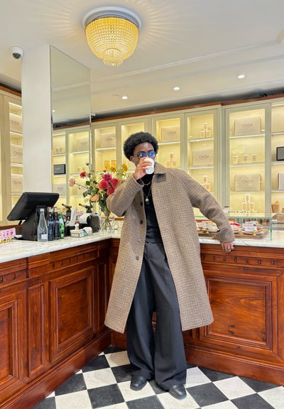 Persona con un abrigo largo de cuadros y gafas azules bebe de una taza, apoyada en el mostrador de madera de una cafetería con flores y botellas de bebida.