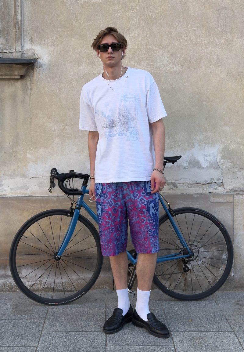 Jeune homme portant des lunettes de soleil, un t-shirt blanc, un short à motifs et des mocassins se tient à côté d’un vélo de route bleu contre un mur beige.