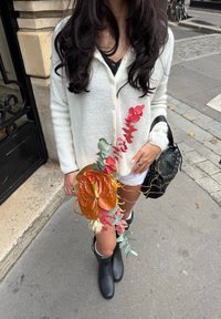 Valge kudum kardigan musta pluusi kohal, hoides kätt oranži ananthuriumi, roosa ja rohelise lehestikuga kimbu. Mustad kummisaapad tänaval.