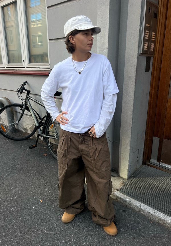 Persona con gorra blanca, camisa blanca de manga larga y pantalones marrones holgados se encuentra en una calle cerca de una bicicleta y la entrada de un edificio.