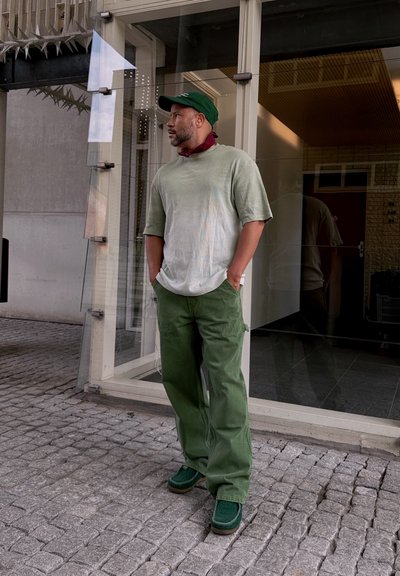 Camiseta grande de color verde con un tono más claro, pantalones cargo verdes, zapatos verdes y una gorra verde oscura. Notable bandana roja alrededor del cuello.