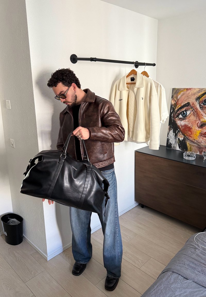 Homme portant une veste en cuir marron et un jean, tenant un grand sac de sport noir à l'intérieur d'une pièce avec des vêtements suspendus et une peinture abstraite de visage.