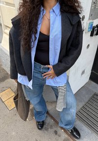 Casaco oversized em mistura de lã preta, camisa de botões azul clara, top preto, jeans de denim azul desgastados, sapatos pretos, detalhe em tecido listrado.