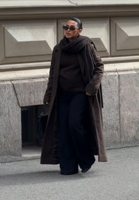 Palton lung maro peste un pulover închis la culoare și pantaloni negri, accesorizat cu ochelari de soare negri și o geantă neagră. Materiale texturate.