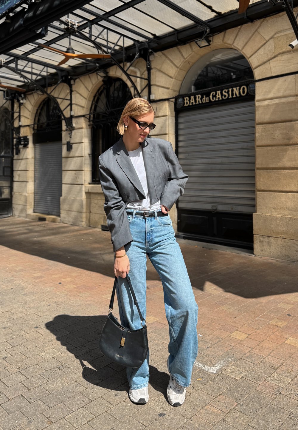Blazer grigio corto, t-shirt bianca, jeans a gamba larga blu chiaro, borsa nera e sneaker bianche spesse su pavimentazione in cobblestone.