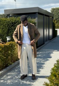 Καφέ διπλό γυναικείο παλτό πάνω από ένα λευκό μπλουζάκι, συνδυασμένο με φαρδιά λευκά παντελόνια και καφέ παπούτσια. Πράσινοι θάμνοι και ένα κτήριο στο παρασκήνιο.