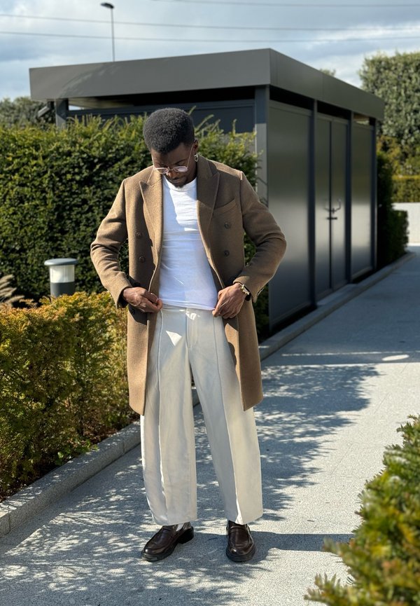 Brown double-breasted coat over a white t-shirt, paired with loose white trousers and brown shoes. Green shrubs and a building in the background.