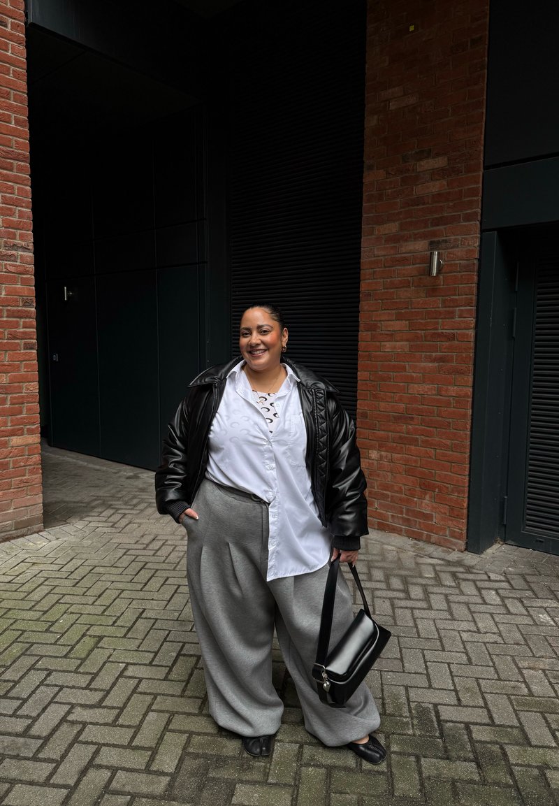 Personne souriante portant une veste en cuir noire, une chemise blanche, un pantalon large gris, tenant un sac à main noir, debout sur un sol pavé près d'un mur en briques.