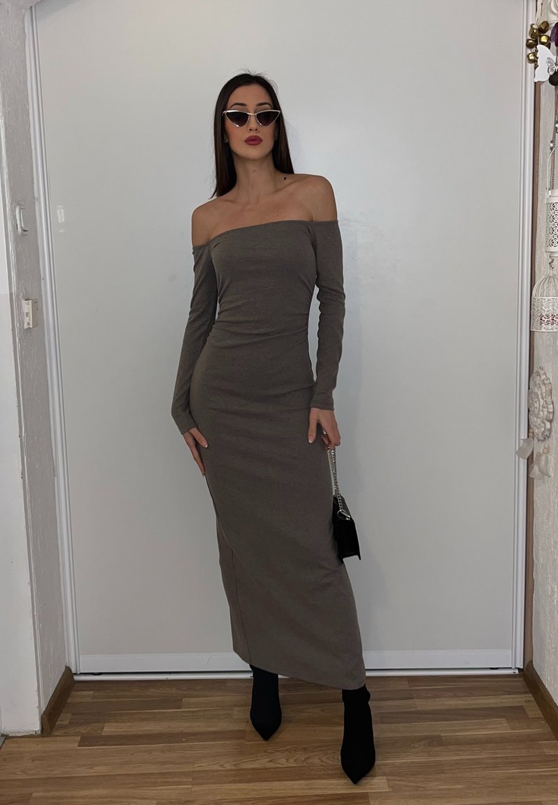 Femme portant une longue robe marron à manches tombantes, des bottes noires, des lunettes de soleil œil de chat, et tenant un petit sac à main noir à l'intérieur.