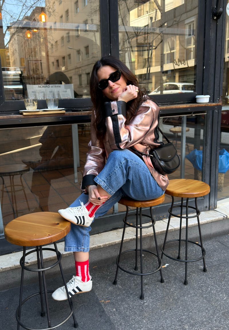 Femme portant des lunettes de soleil, une veste rose, un jean bleu, des baskets blanches et des chaussettes rouges assise sur un tabouret en bois à l'extérieur d'un café avec un sac à main noir.