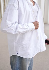 Biela košeľa s button-down golierom a modrým logom na leme, spárovaná so svetlomodrými džínsami. Košeľa má voľný strih a pokrčenú textúru.