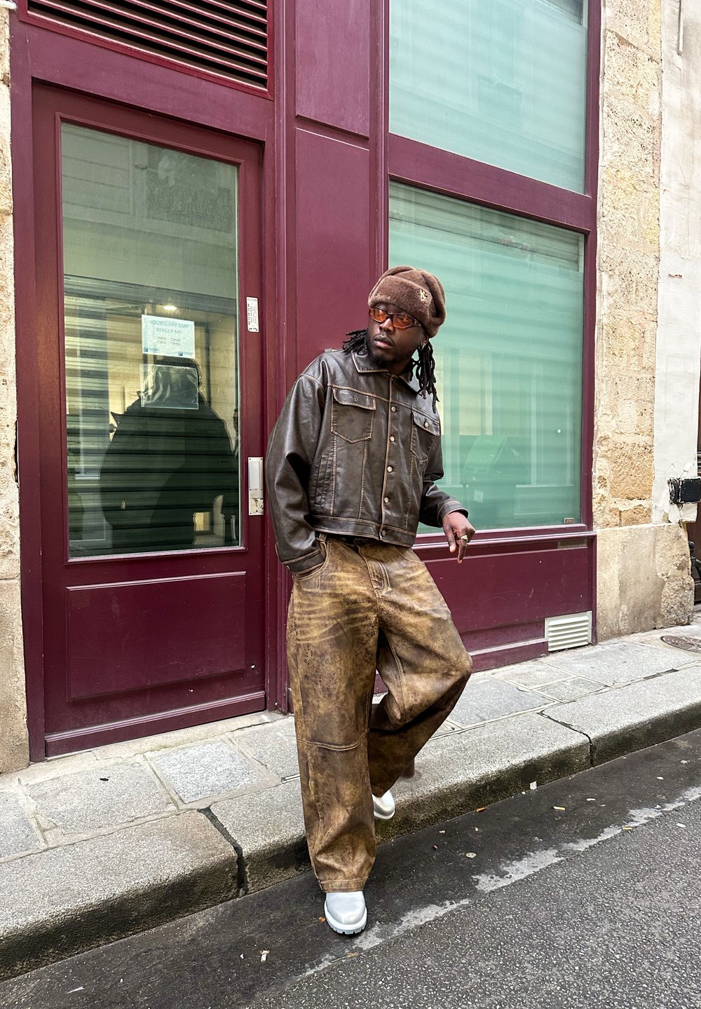 Καφέ δερμάτινο μπουφάν με cropped σχέδιο, oversized καφέ παντελόνια με υφή και μπεζ παπούτσια, μπροστά από ένα μπορντό τοίχο.