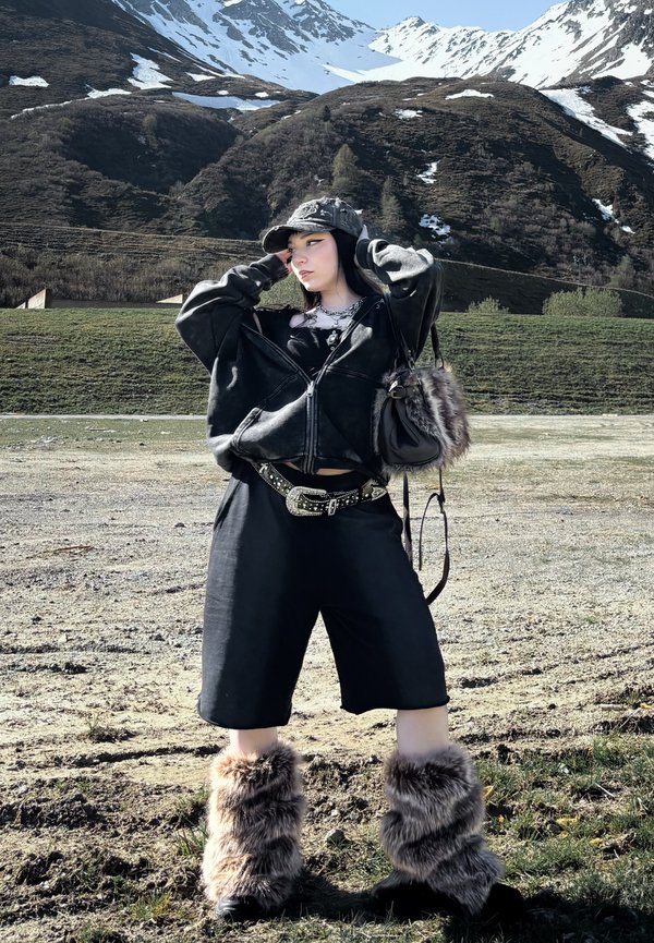 En ung person iført en sort oversized jakke, hat, shorts og pelsede støvler poserede udendørs med sneklædte bjerge og grønne bakker i baggrunden.