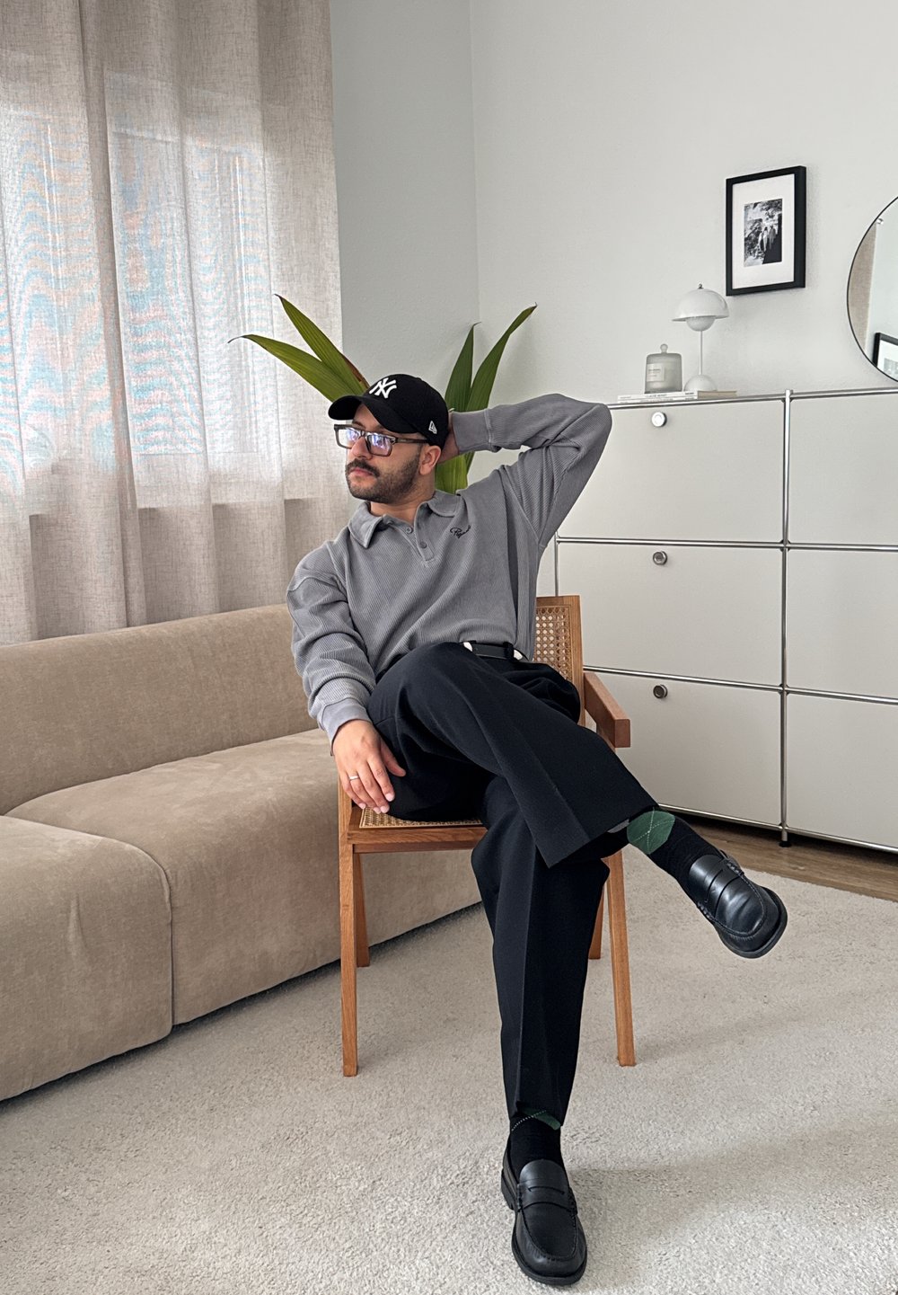 Grå ribbet polo skjorte, sorte bukser, sorte loafers og sokker med grønne accenter. Træstol, beige sofa, potteplante og minimal dekoration.