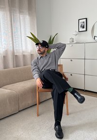 Grå ribbet polo skjorte, sorte bukser, sorte loafers og sokker med grønne accenter. Træstol, beige sofa, potteplante og minimal dekoration.