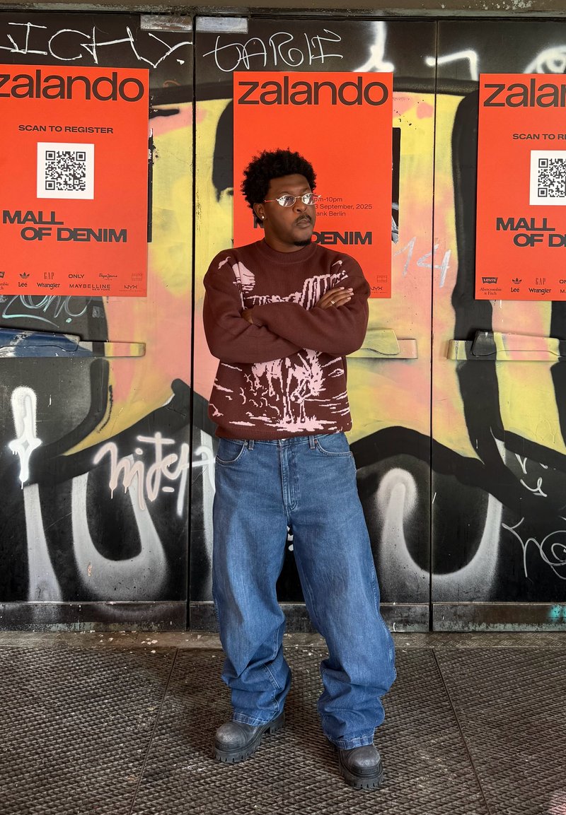Pull en maille bordeaux avec motif graphique rose, jeans baggy bleus et bottes noires épaisses, sur fond de graffiti coloré et d'affiches promotionnelles.