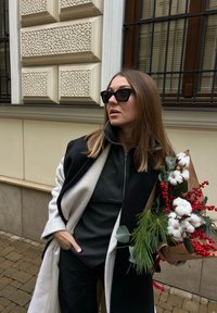 Femme en manteau noir et blanc portant des lunettes de soleil, tenant un bouquet de coton, de baies rouges et de branches de pin vertes en plein air.